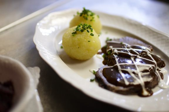 Sauerbraten mit Kloß auf einem weißen Teller, daneben eine Schale mit Rotkraut und ein Glas Wein.